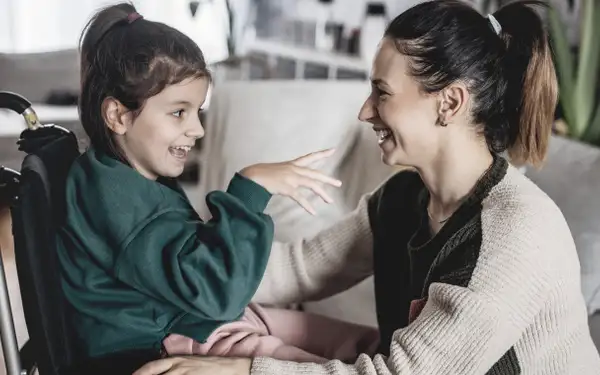 Eine junge Frau, die ein duales Studium in Sozialer Arbeit absolviert, interagiert lächelnd mit einem lachenden Mädchen im Rollstuhl.