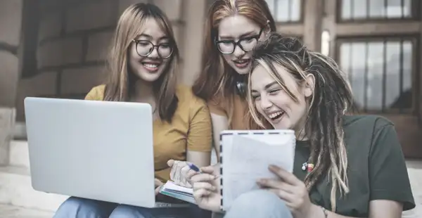 Drei Studentinnen, die Gender Studies studieren, arbeiten gemeinsam an einem Projekt. Sie lachen und diskutieren, während sie einen Laptop und Notizbücher verwenden.