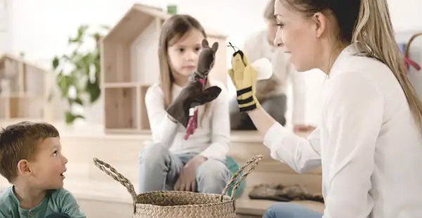 Eine junge Frau, die eine Weiterbildung in der Erziehung macht, spielt mit Handpuppen vor zwei aufmerksam zuschauenden Kindern.