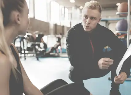Sportpsychologe kniet neben Sportlerin im Fitnessraum und erklärt ihr etwas