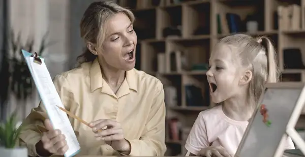 Eine junge Frau, die ein Fernstudium in Logopädie absolviert, übt Ausspracheübungen mit einem kleinen Mädchen.