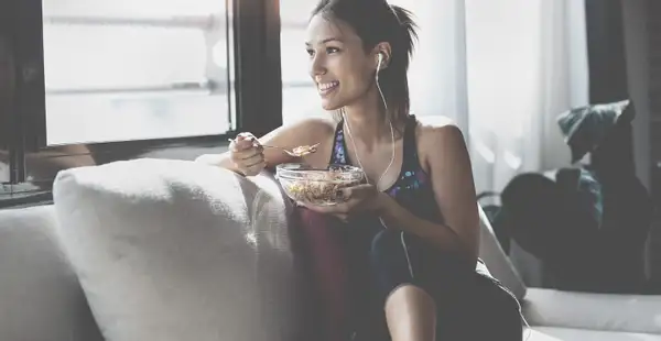 Sportlerin sitzt in Sportbekleidung auf dem Sofa und isst Müsli