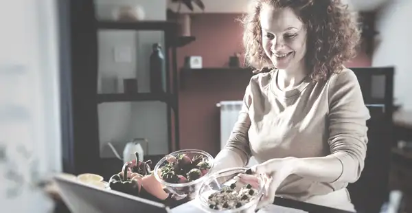 Eine Frau, die ein Fernstudium in Ernährungswissenschaften absolviert, steht mit einem Lächeln auf den Lippen in der Küche, vor sich zwei Schüsseln mit Lebensmitteln. 