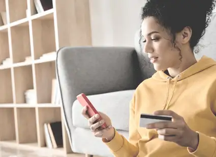 Junge Frau in einem sonnengelben Pullover sitzt mit Kreidtkarte und Smartphone auf einer Couch.