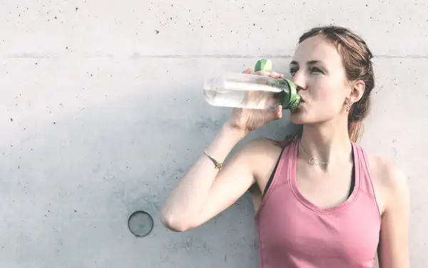 Junge Sportlerin trink an eine Wand gelehnt lächelnd Wasser.