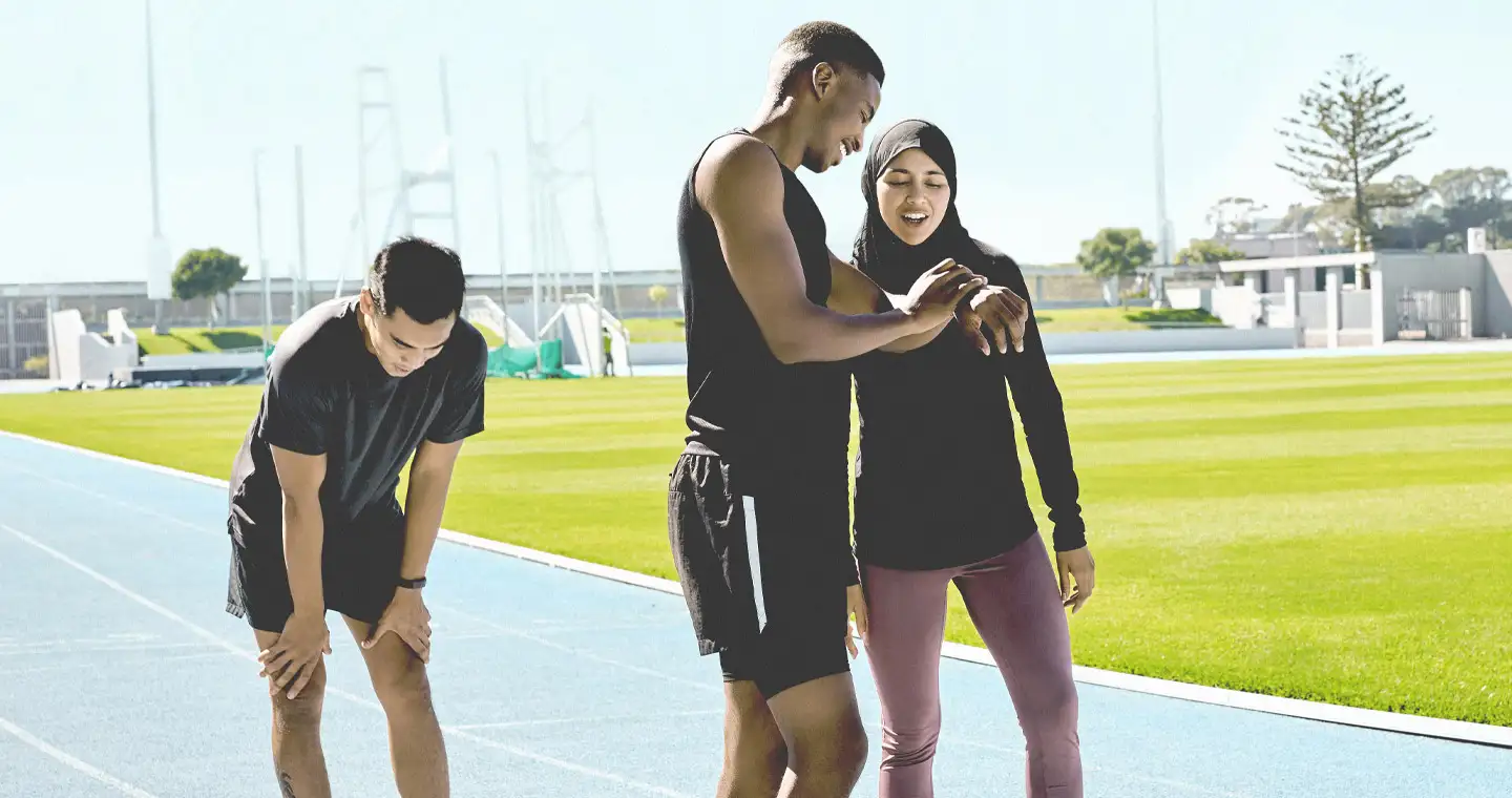 Drei angehende Sportstudierende trainieren für den Lauf auf Zeit der Sporteignungsprüfung. Sie stehen auf der Laufbahn und kontrollieren ihre Laufzeit auf der Armbanduhr.
