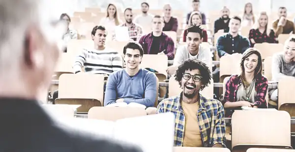 In einem Hörsaal sitzen lächelnde Studierende und hören einem Professor zu. 