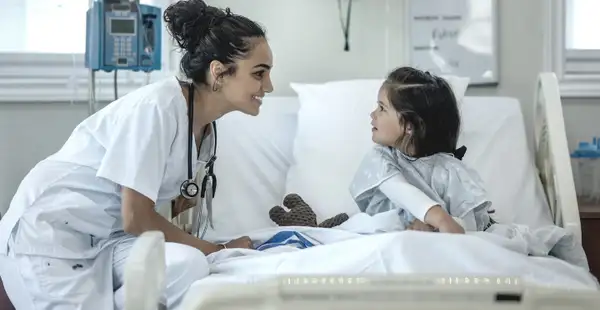 Eine junge Frau, die ein Fernstudium in der Pflege absolviert, spricht lächelnd mit einem kleinen Mädchen, das in einem Krankenhausbett liegt. 