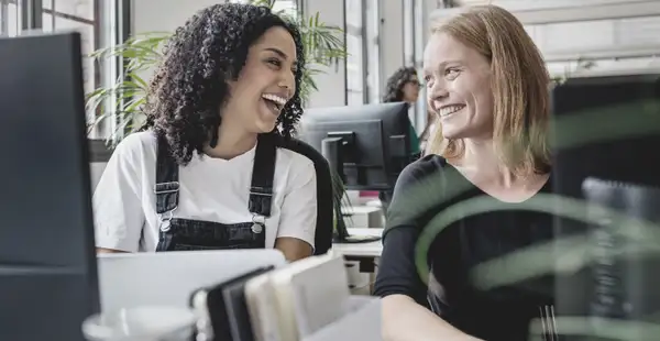 Zwei Masterstudentinnen im Studiengang Public Management arbeiten lächelnd in der Bibliothek an einem gemeinsamen Projekt.