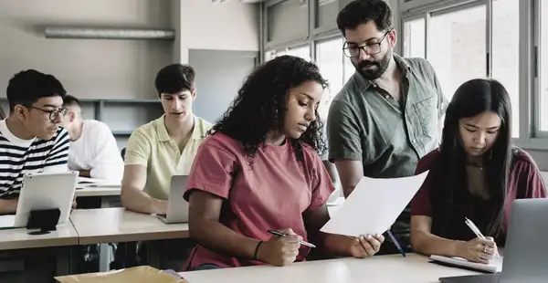 Ein Mann, der Berufspädagogik studiert hat, unterstützt eine Gruppe von Studierenden in einem Klassenzimmer. Alle sind konzentriert und arbeiten an Laptops.