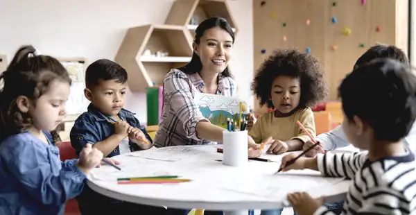 Eine Sozialpädagogin sitzt an einem runden Tisch und malt zusammen mit einer Gruppe von Kindern.