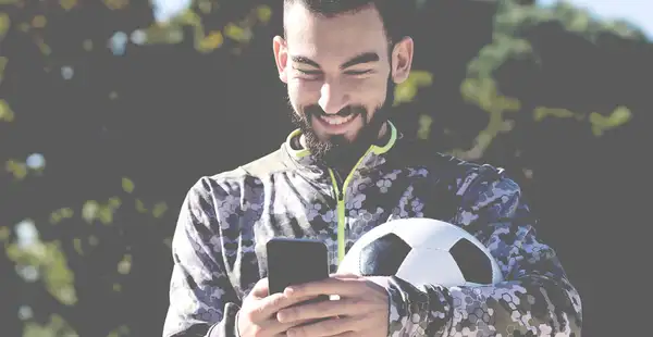 Ein Sportdirektor in Fußball hat einen Fußball unter den Arm geklemmt und tippt an seinem Smartphone.