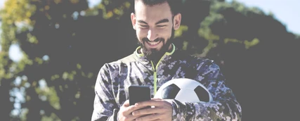 Ein Sportdirektor in Fußball hat einen Fußball unter den Arm geklemmt und tippt an seinem Smartphone.