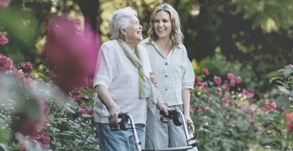 Eine Studentin der Sozialen Gerontologie begleitet eine ältere Frau mit einem Rollator auf einem Spaziergang durch einen Garten.