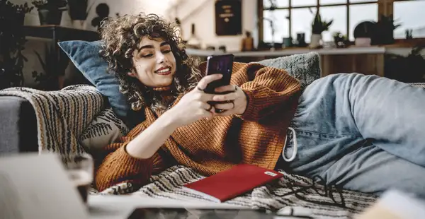 Eine junge Studentin liegt mit einem Buch und ihrem Handy in den Händen auf der Couch.