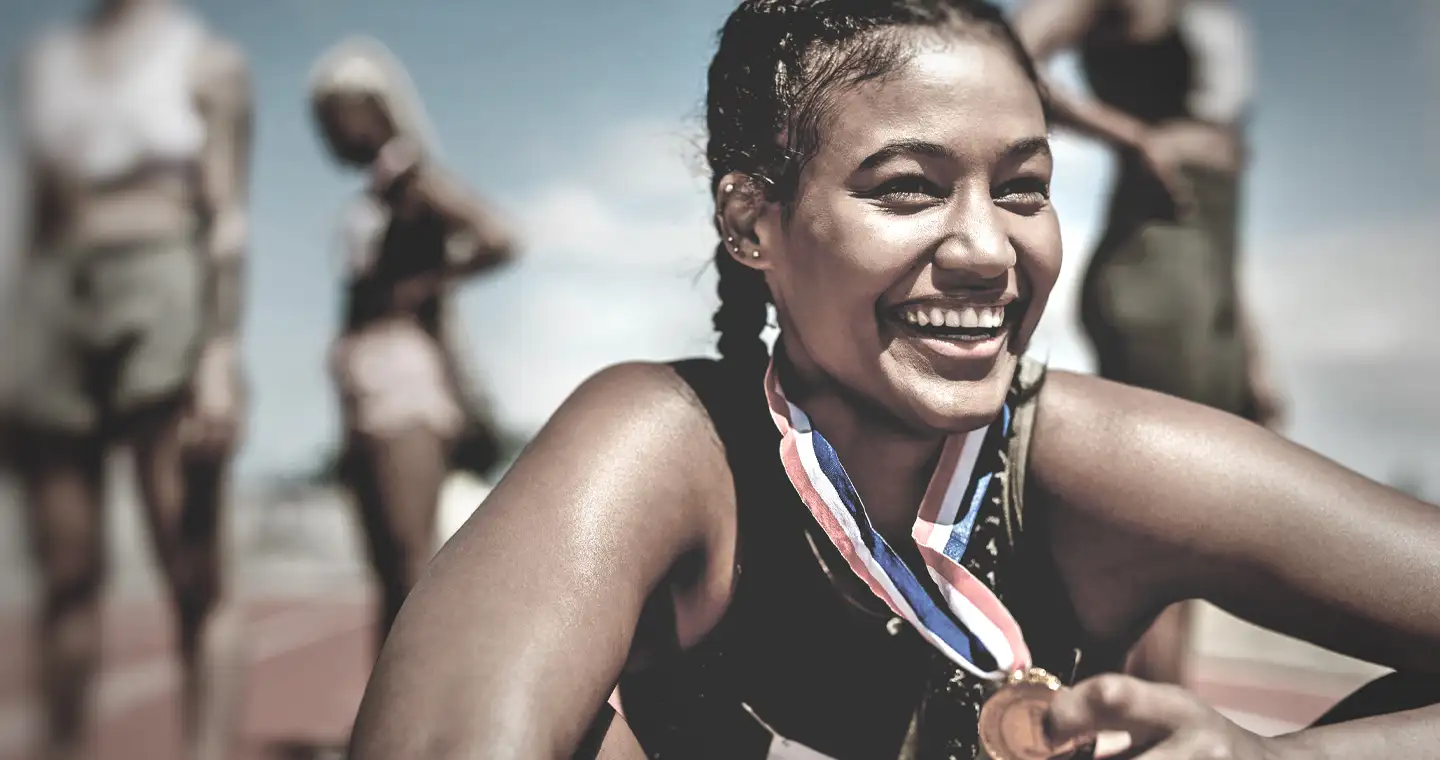 Eine Sportler*in sitzt nach einem Wettlauf erschöpft aber gut gelaunt auf dem Laufplatz und trägt ihre Siegermedaille um den Hals.