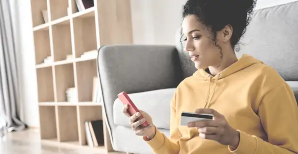 Junge Frau in einem sonnengelben Pullover sitzt mit Kreidtkarte und Smartphone auf einer Couch.