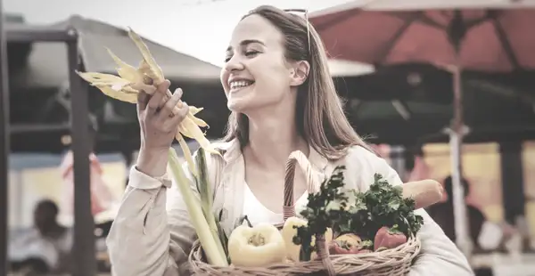 Eine Ernährungsberaterin in IHK-Ausbildung schlendert mit einem Korb voller Einkäufe in der Hand lächelnd über einen Gemüsemarkt.