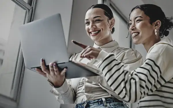 Zwei Medienmanagerinnen stehen im Büro und schauen gut gelaunt auf den Laptop, den eine der Frauen trägt.